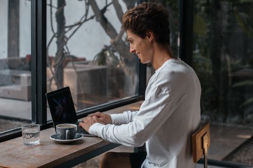 Free A young man in a white long sleeve shirt smiles while typing on his laptop at a coffee shop. Stock Photo