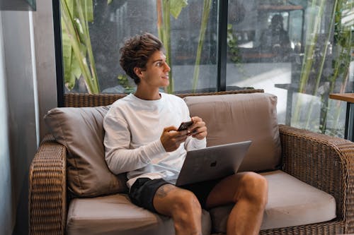 Free Young Man Sitting on a Couch Using Laptop and Phone Smiling  Stock Photo