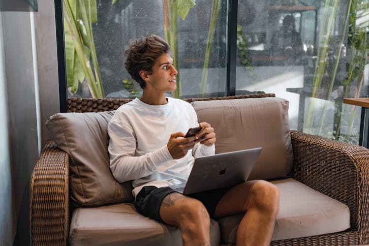 Young Man Sitting On A Couch Using Laptop And Phone Smiling 