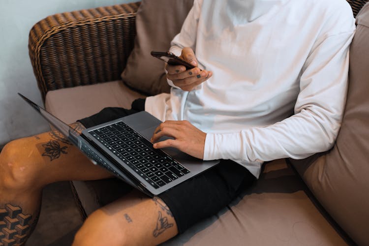 Man With Tattoos Sitting On A Couch And Using Laptop And Phone 