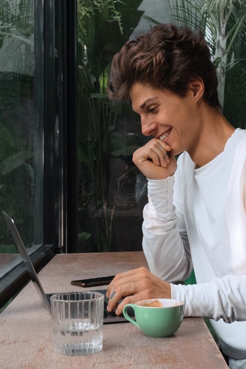 Free A young man with short brown hair smiling at a laptop in a café with a cup of coffee. Stock Photo
