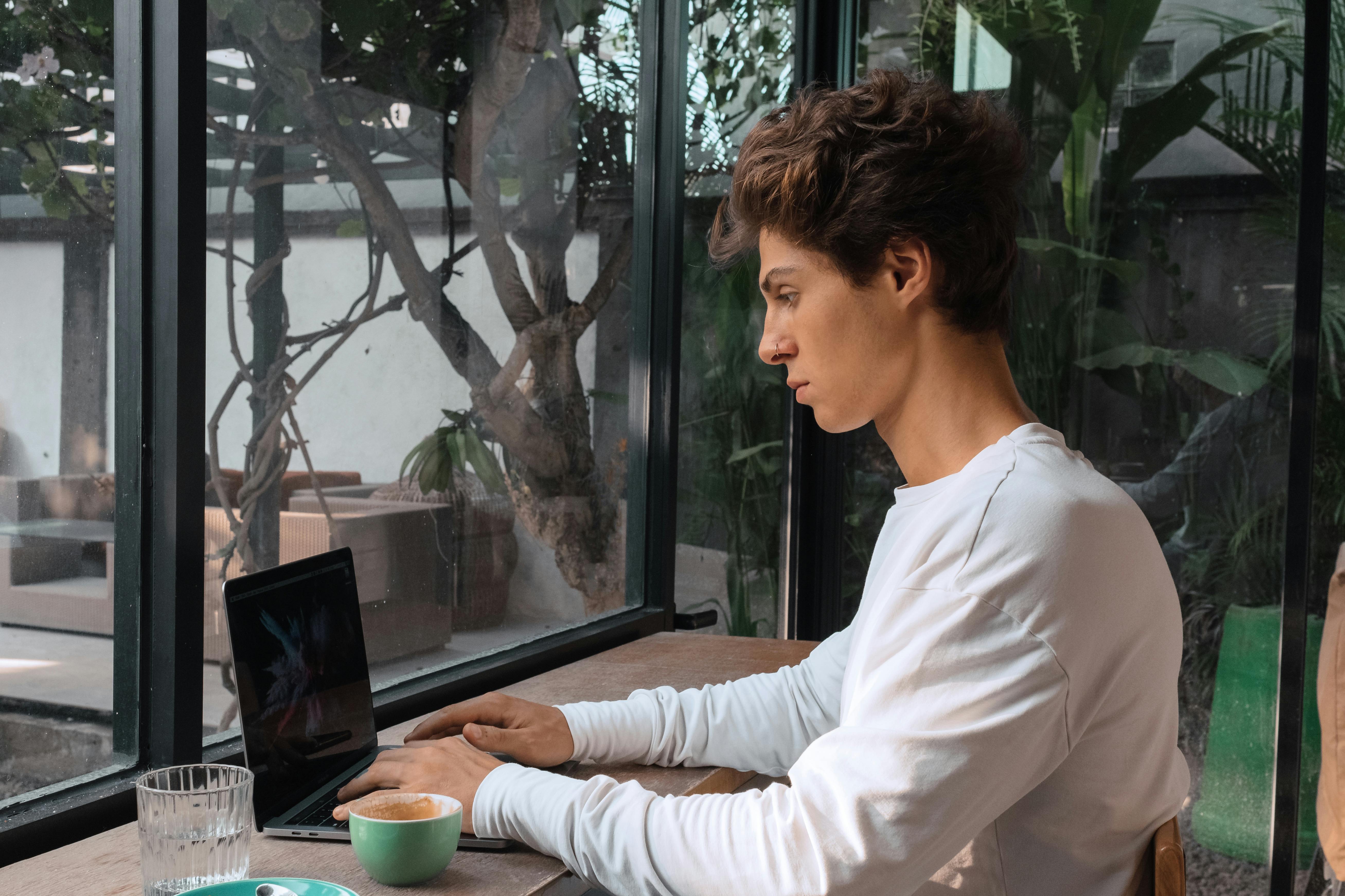 一位专注的年轻人坐在舒适的咖啡馆里，靠着窗边，一边喝着咖啡一边用笔记本电脑打字