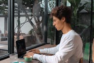 一位专注的年轻人坐在舒适的咖啡馆里，靠着窗边，一边喝着咖啡一边用笔记本电脑打字