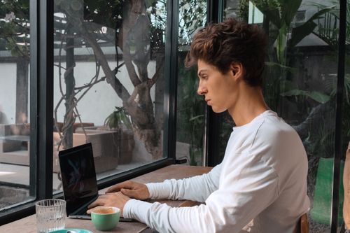 Free Focused young man typing on a laptop inside a cozy café, enjoying coffee by the window. Stock Photo