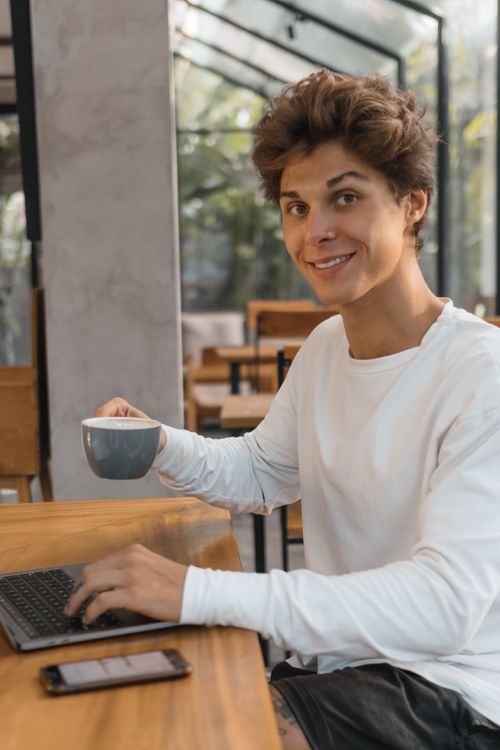 Free Smiling young man using a laptop and sipping coffee in a stylish, modern café setting. Stock Photo