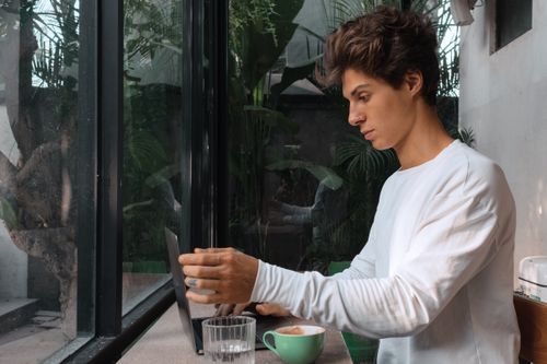 Free A young man working on his laptop in a modern café surrounded by greenery. Stock Photo