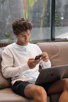 A young man sitting indoors, multitasking with a smartphone and laptop in a relaxed environment.