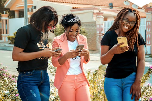 Happy ethnic women in trendy wear text messaging on cellphones on city street