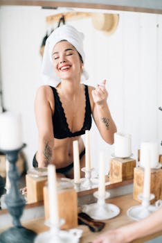 Woman in black lingerie with a towel on her head, smiling joyfully at her reflection in a brightly lit room.