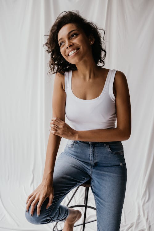 Free Smiling Woman in White Tank Top and Blue Denim Jeans Sitting on Chair Stock Photo