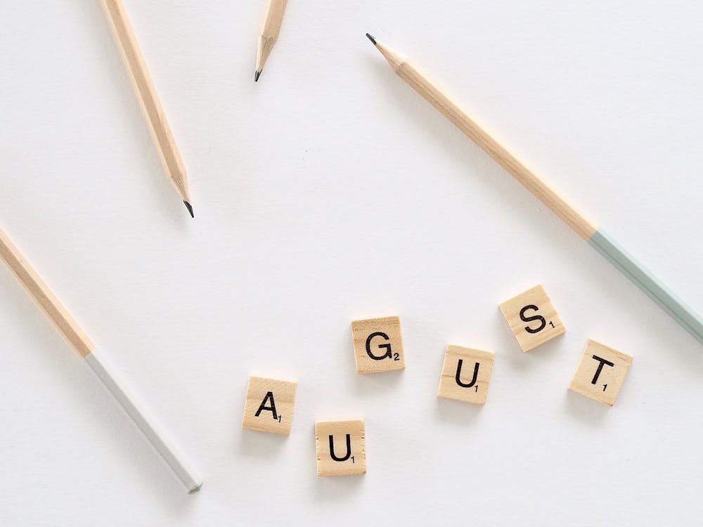 Flat lay of small wooden cubes with letters saying August arranged on white background with scattered pencils on table in room
