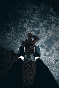 From above of legs and reflection of faceless person in black trousers and sneakers on wet pavement in daylight
