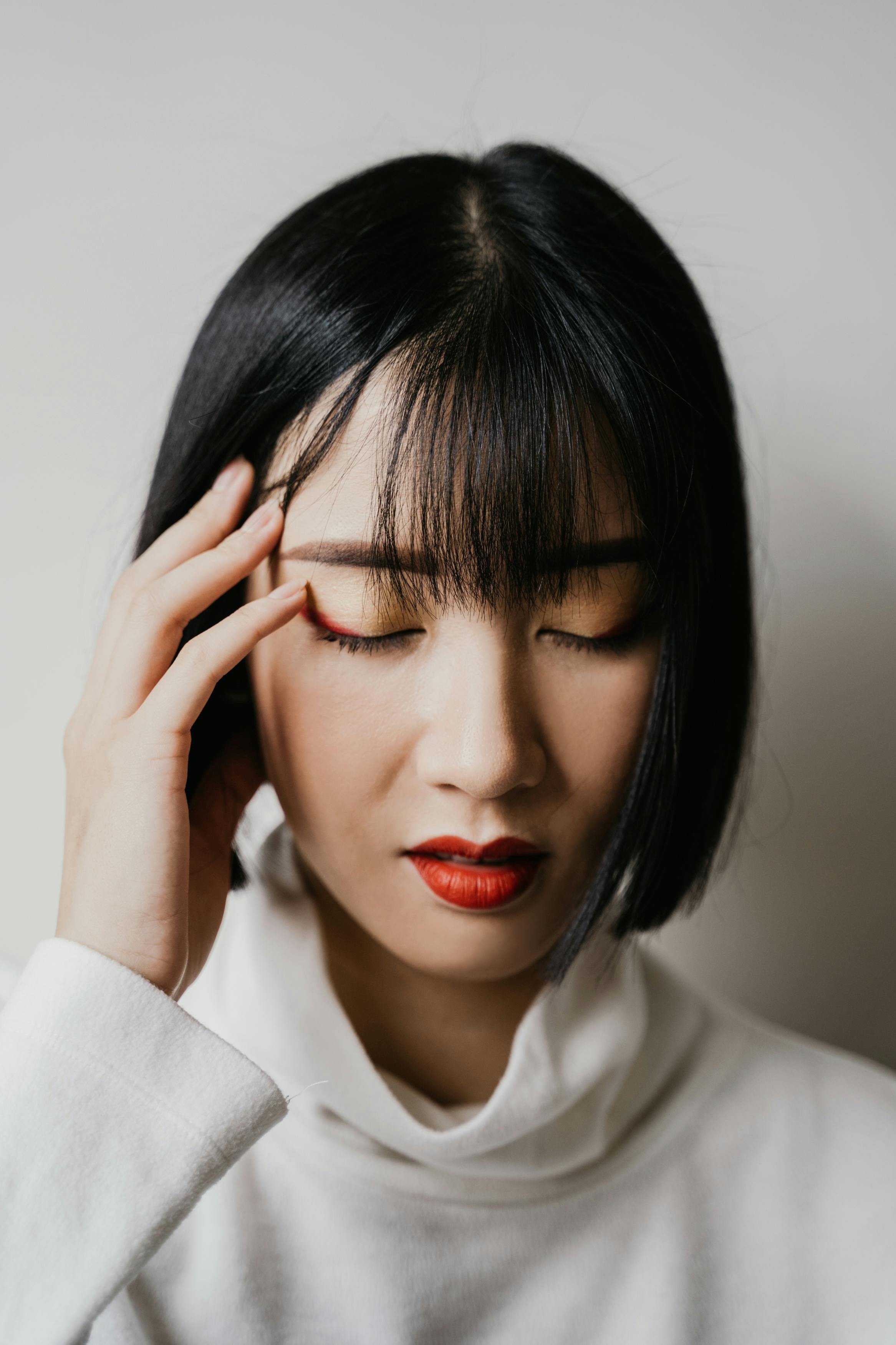 Close-up portrait of an Asian woman with bangs, eyes closed, and red lips, expressing deep thought.