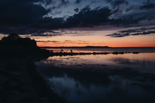 A dramatic sunset reflects on a calm lake surface with silhouetted trees and rocks.
