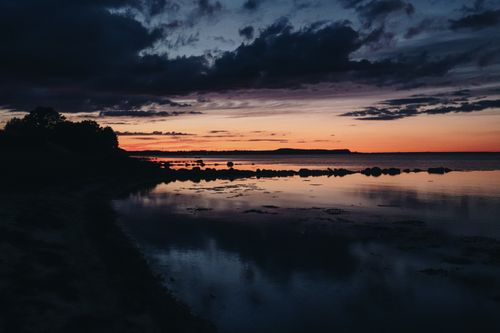 Free A dramatic sunset reflects on a calm lake surface with silhouetted trees and rocks. Stock Photo