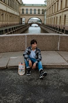 在俄罗斯历史悠久的圣彼得堡，一位年轻的滑板运动员坐在运河旁，背景是标志性的拱门
