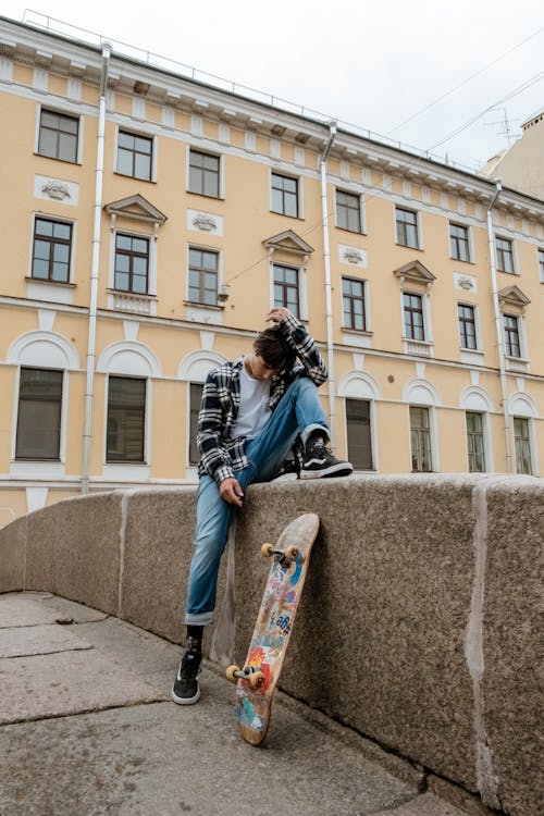 Woman in Black Jacket and Blue Denim Jeans Sitting on Skateboard Near Beige Concrete Building during