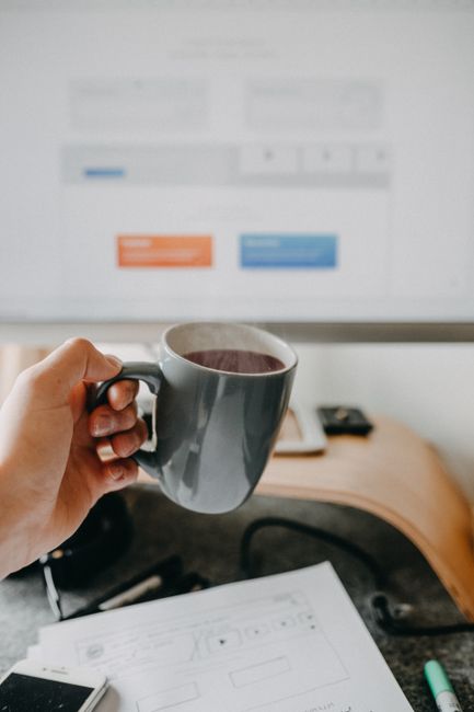 Close-up of a hand holding a coffee mug in front of a computer screen in an office setting, creating a relaxed atmosphere.