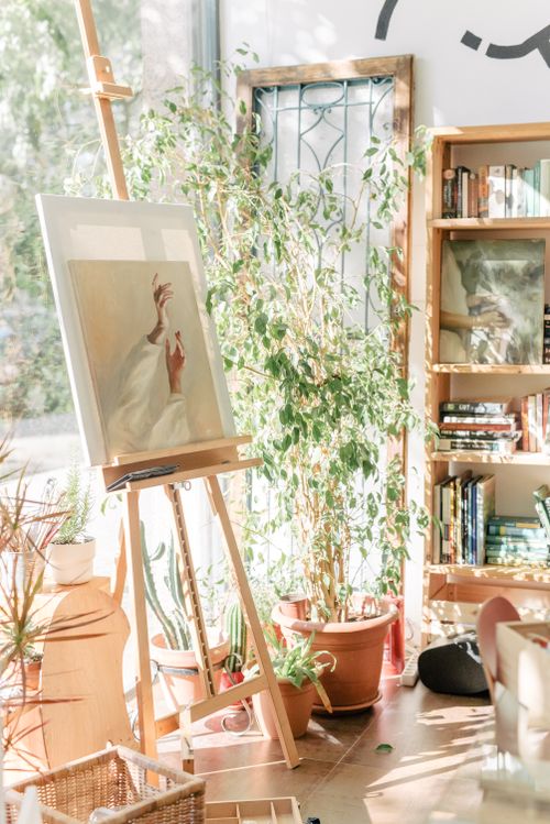 Free A cozy art studio with a canvas on an easel, filled with warm sunlight and surrounded by plants. Stock Photo