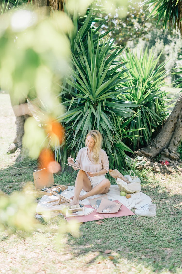 A Woman Sitting On A Picnic Blanket While Sketching