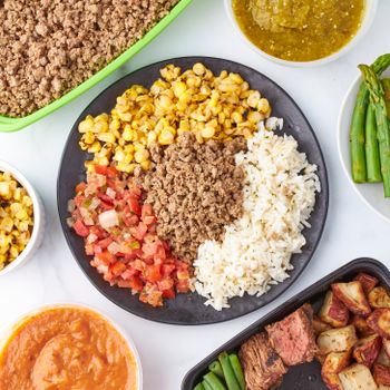A colorful flat lay of a healthy meal featuring rice, corn, asparagus, and more on a white background.