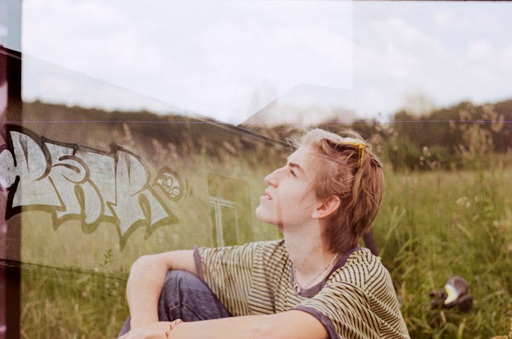 Double exposure of young contemplative male sitting on green meadow under cloudy sky while looking up near house with graffiti