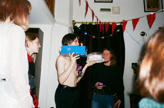 From below of cheerful teen with gift and festive cake standing with naked torso near smiling friends in house with decorative garland