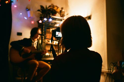 Free Unrecognizable female recording video of friend playing guitar on camera in flat with shiny garlands Stock Photo