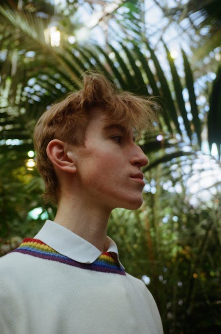 Low angle of young trendy male with contemporary haircut looking forward under lush green tree leaves