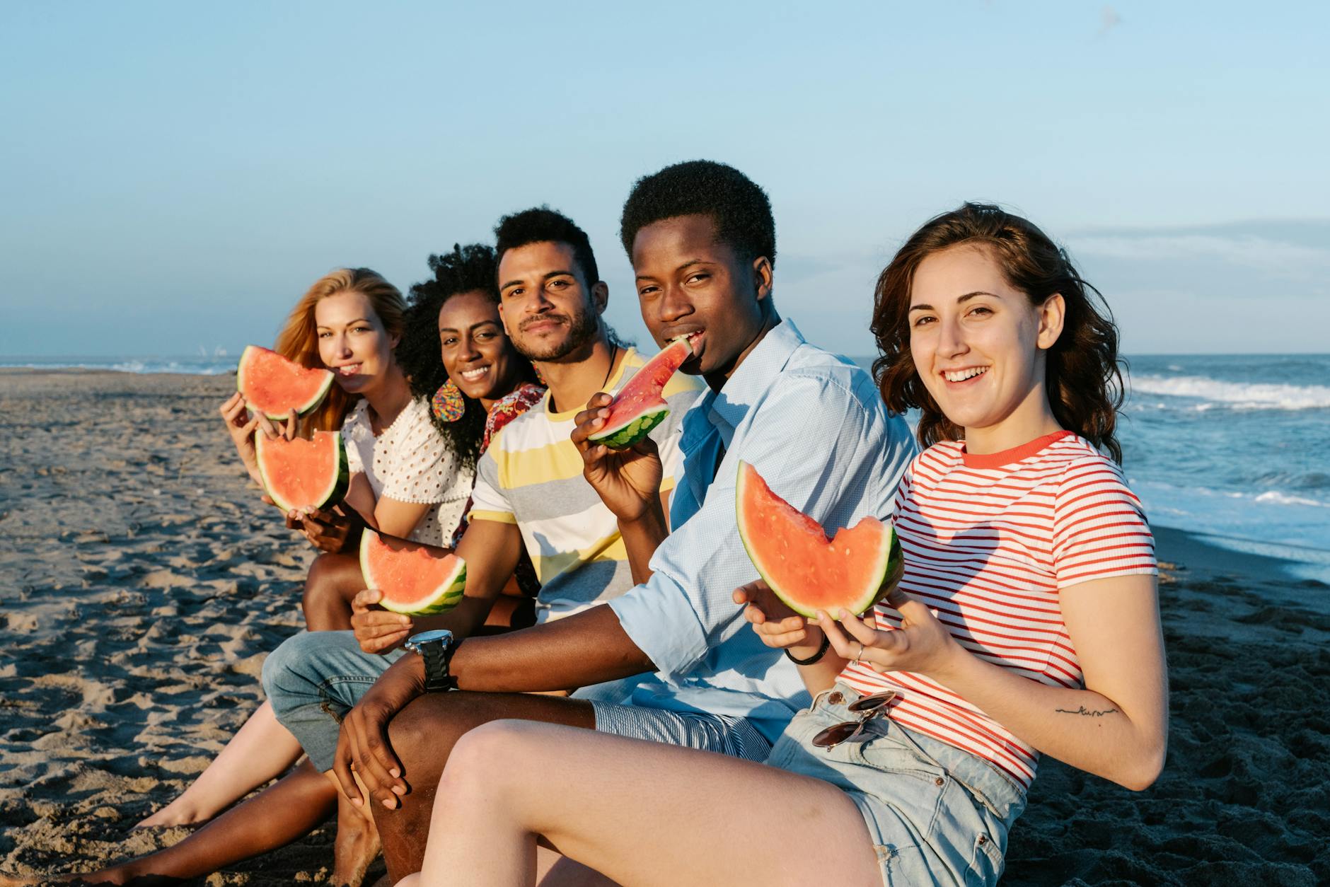 Young content multiracial friends enjoying sweet ripe watermelon slices on sandy sea shore while looking at camera