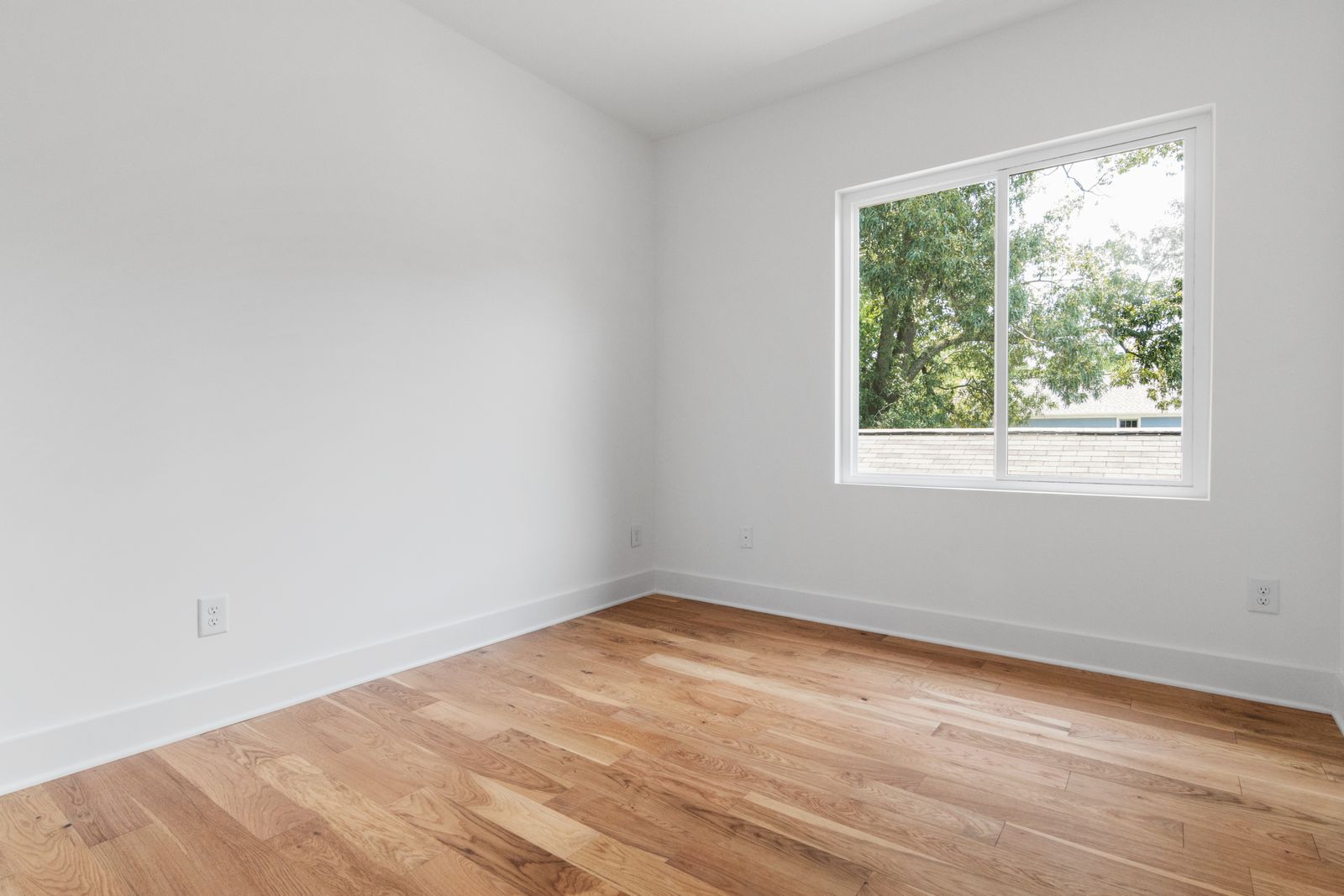 Una habitación vacía con paredes blancas, suelo de madera y una única ventana que da a unos árboles