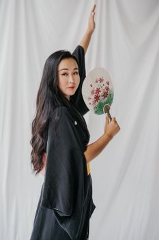 一名身穿黑色和服的女子手持花卉扇子，在白色背景下优雅地摆着姿势