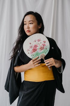Portrait of a woman in a black kimono holding a floral hand fan against a white background.