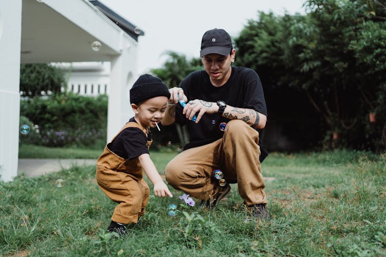 Man In Brown Pants And Boy In Brown Pants And Black Hat Sitting On Green Grass Field And Blowing Soap Bubbles