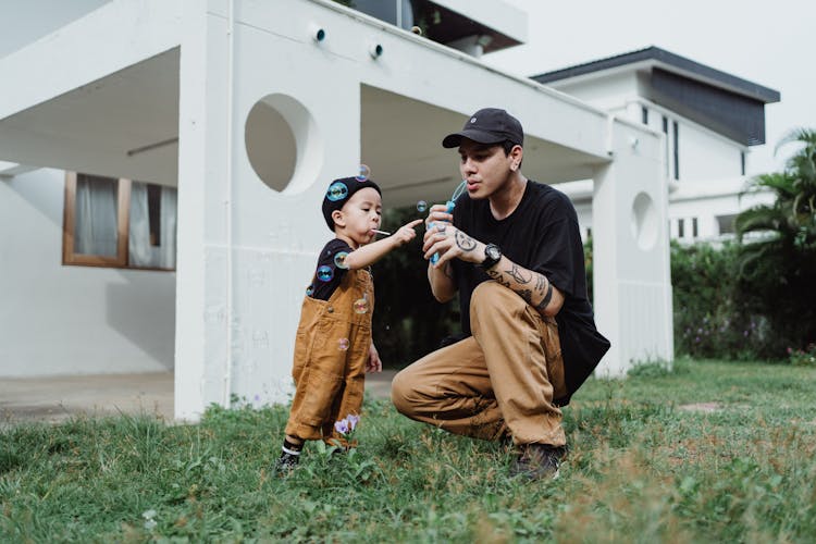 Man In Brown Pants And Boy In Brown Overall Sitting On Grass And Blowing Bubbles