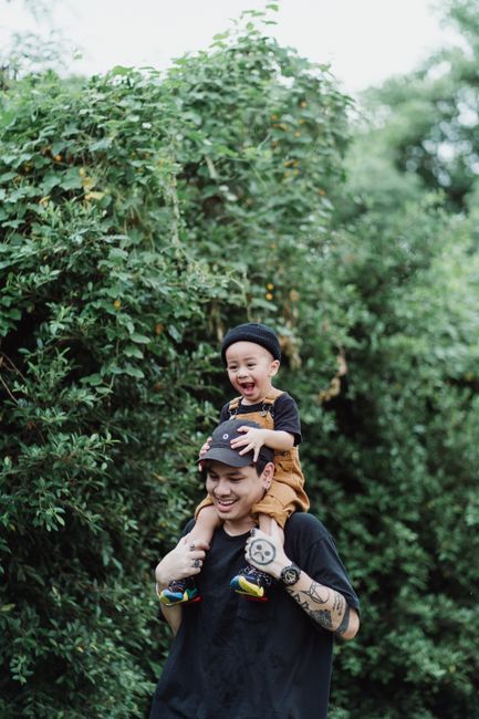 A father carries his laughing son on shoulders in a lush green outdoor setting. Captured in summer.