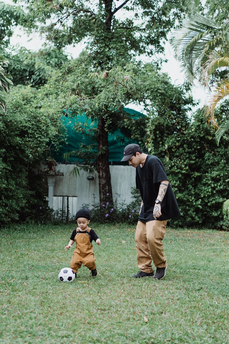 Man In Black T-Shirt And Brown Pants Playing With Boy In Brown Overall And Soccer Ball