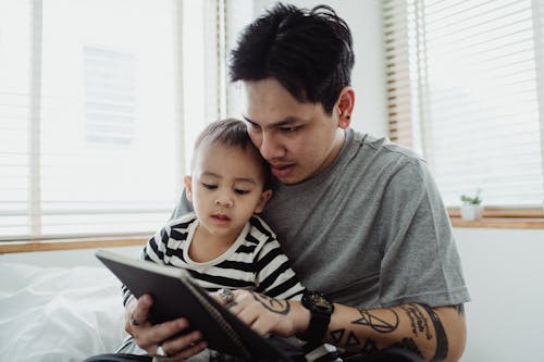 Man in Gray T-Shirt Holding Boy in Striped Long Sleeve Shirt and Using Tablet