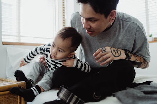 Man in Gray T-Shirt Sitting on Bed Beside Boy in Striped Long Sleeve Shirt