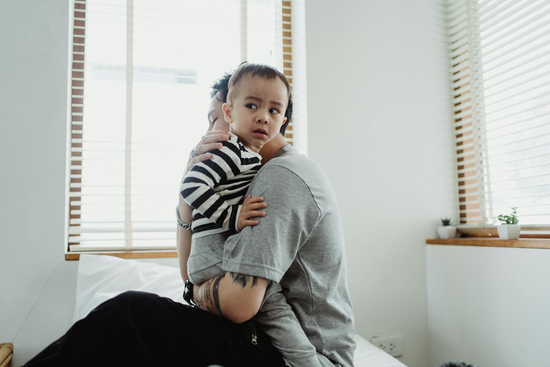 Father holding and comforting his young son by the window, showcasing warmth and family bond.