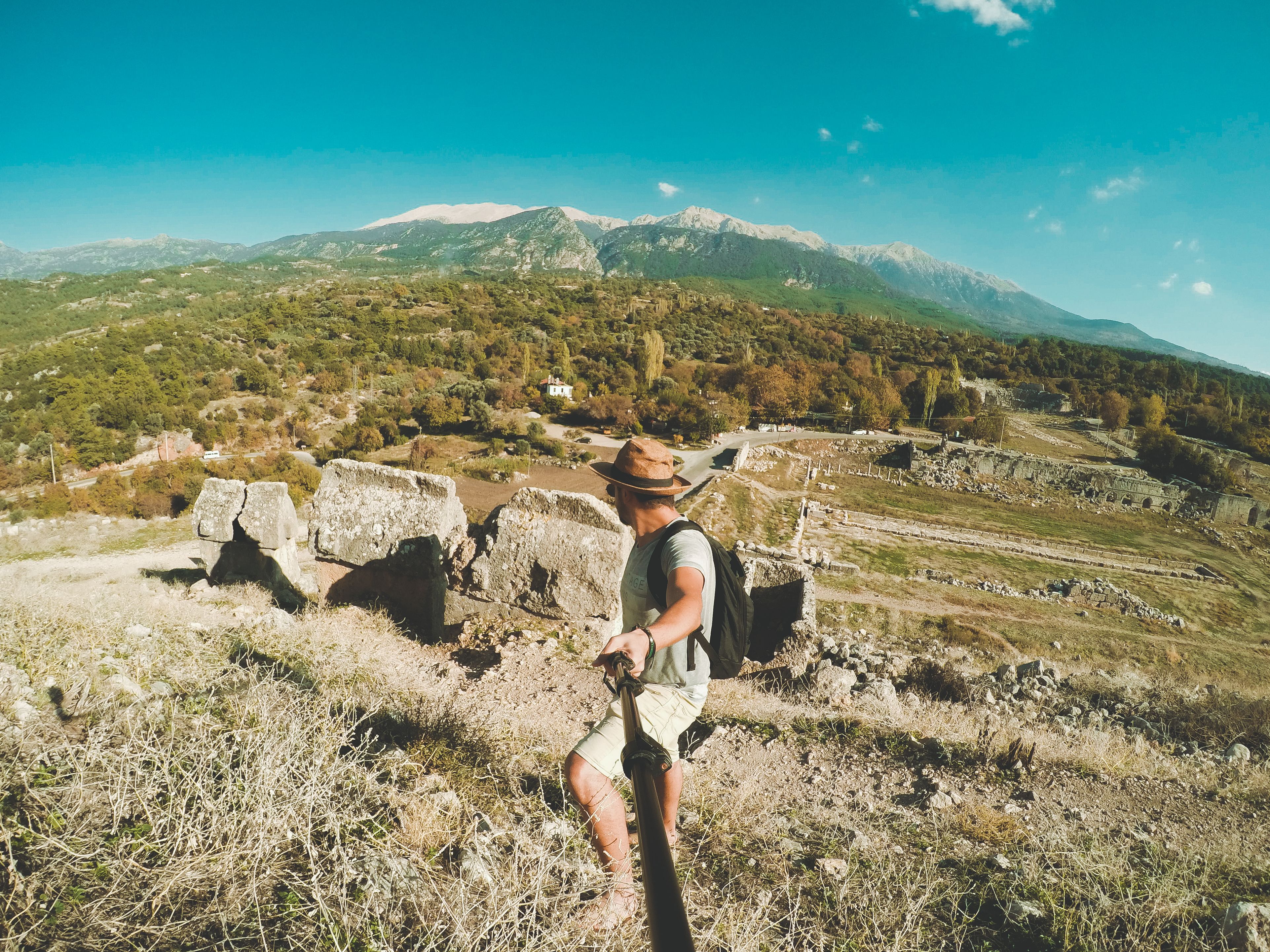 Traveler with backpack explores ancient ruins in a mountainous landscape under a bright blue sky.