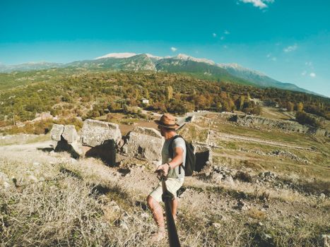 Traveler with backpack explores ancient ruins in a mountainous landscape under a bright blue sky.