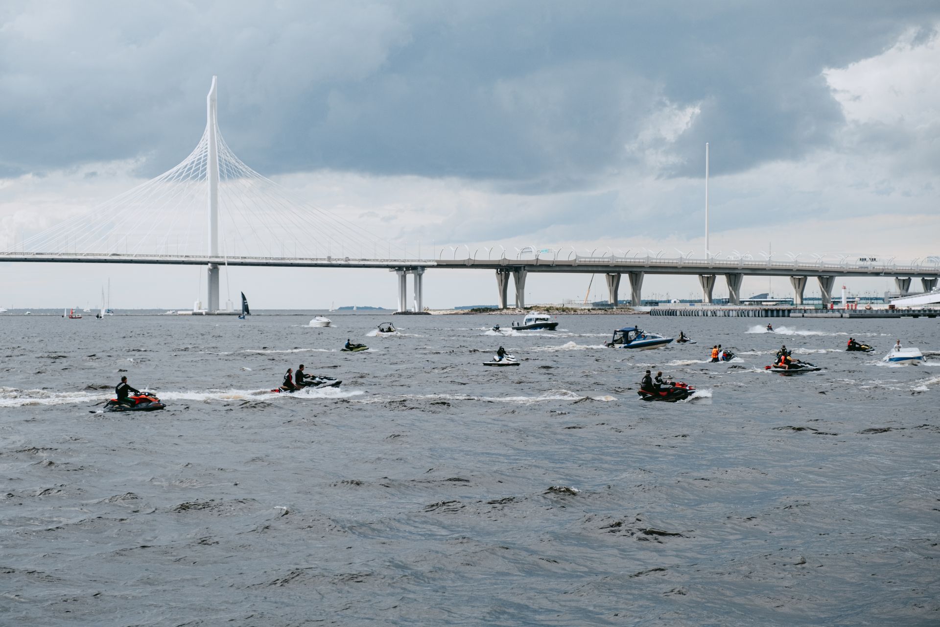 Multiple jet skis and boats navigating water with a bridge in the background under a cloudy sky.
