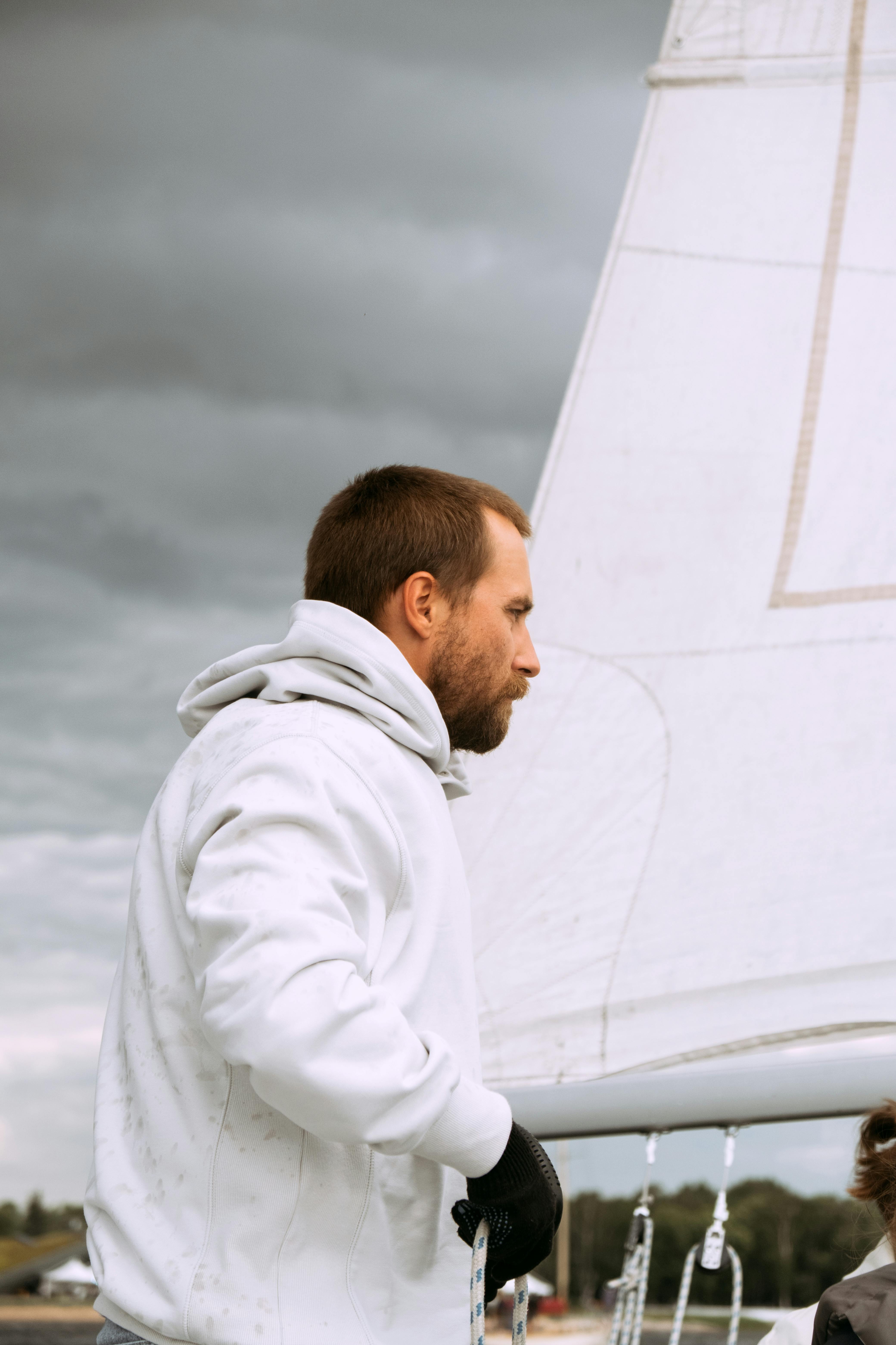 A man in a white hoodie navigates a yacht under overcast skies, epitomizing adventure.