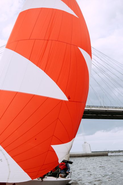 Dynamic shot of a sailboat with a striking red sail navigating beneath a modern cable-stayed bridge.