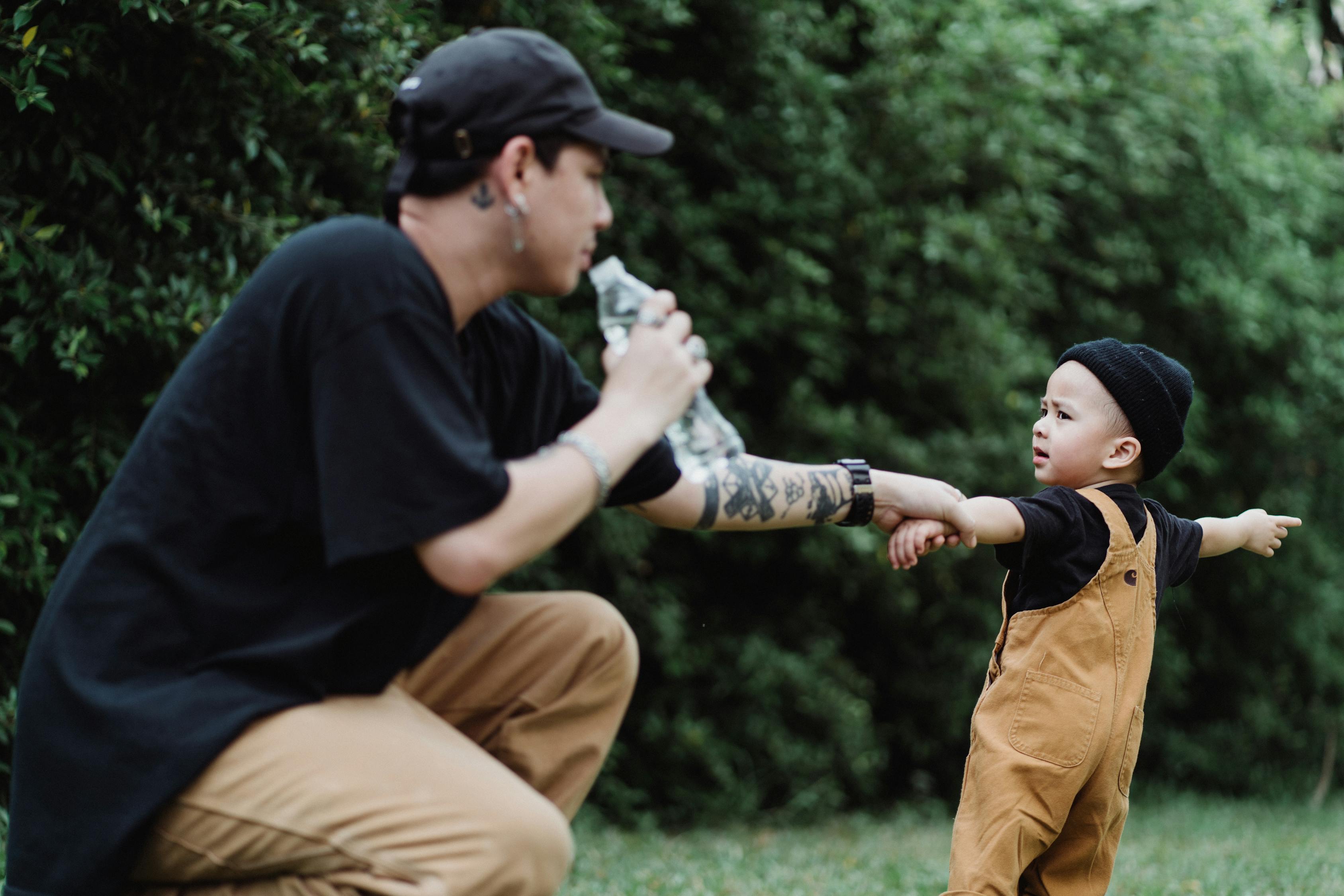 Man in Black T-shirt and Brown Pants Holding Boy's Hand While Drinking Water
