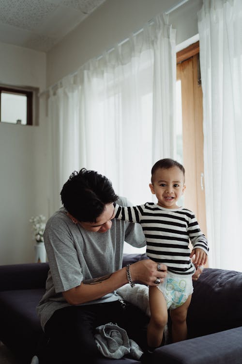 Little Boy in Striped Shirt and Diaper Standing on the Couch Beside Man in Gray T-Shirt