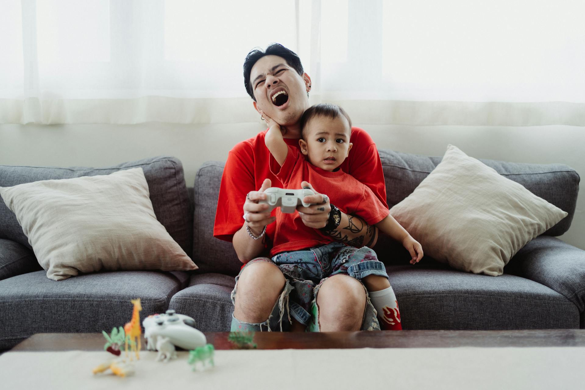 Father and son sharing a playful moment gaming on the sofa with toys scattered around.