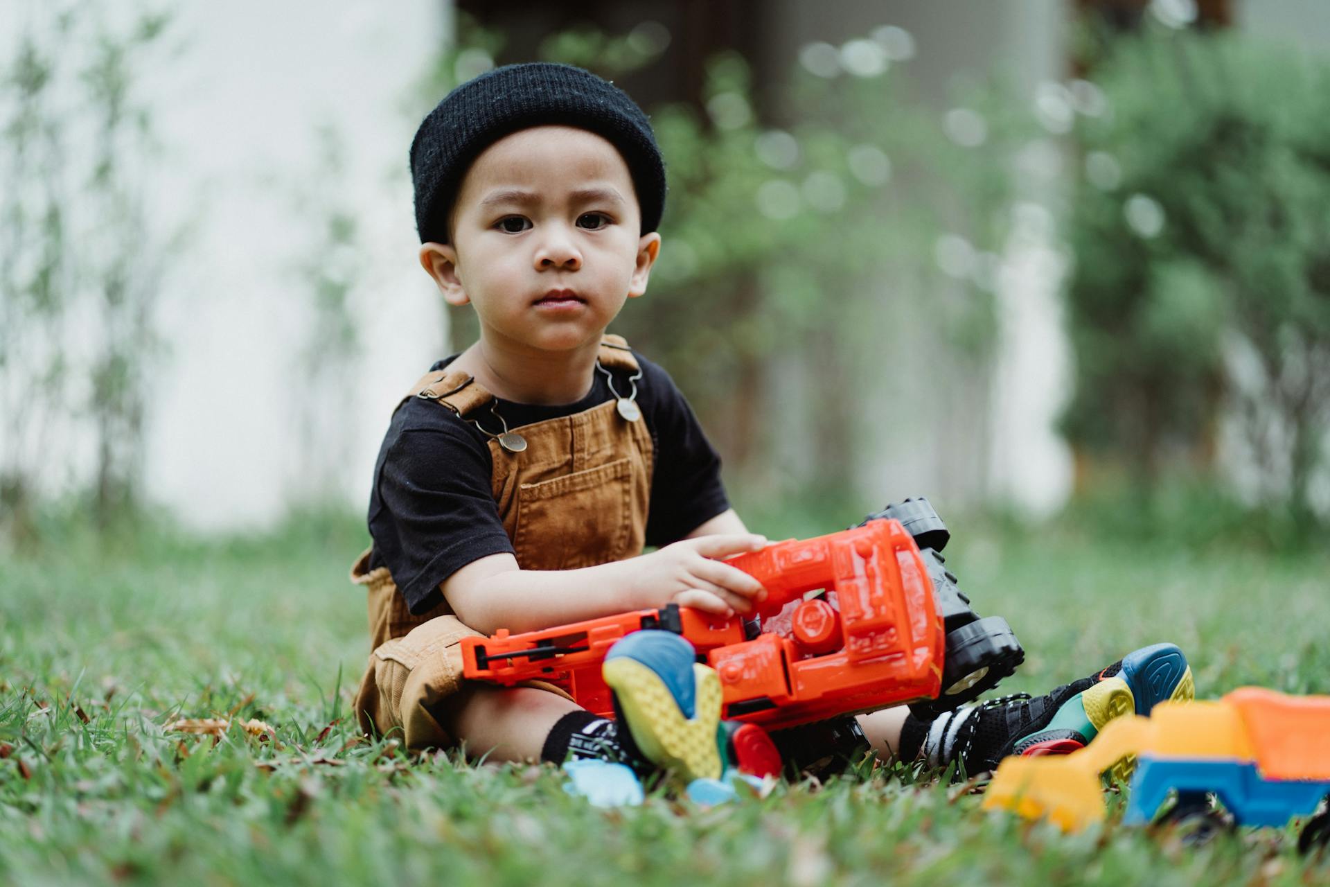 A young boy in overalls and a beanie plays with a toy truck on grass.