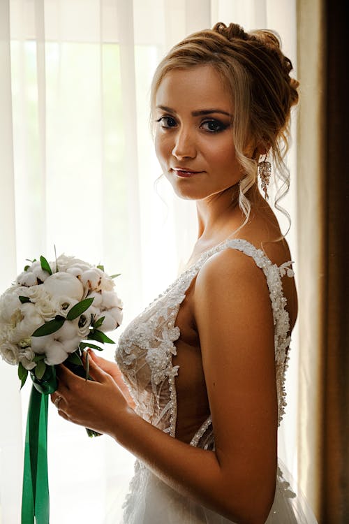 Side view of dreamy young bride in elegant white dress standing near window with white bouquet and looking at camera